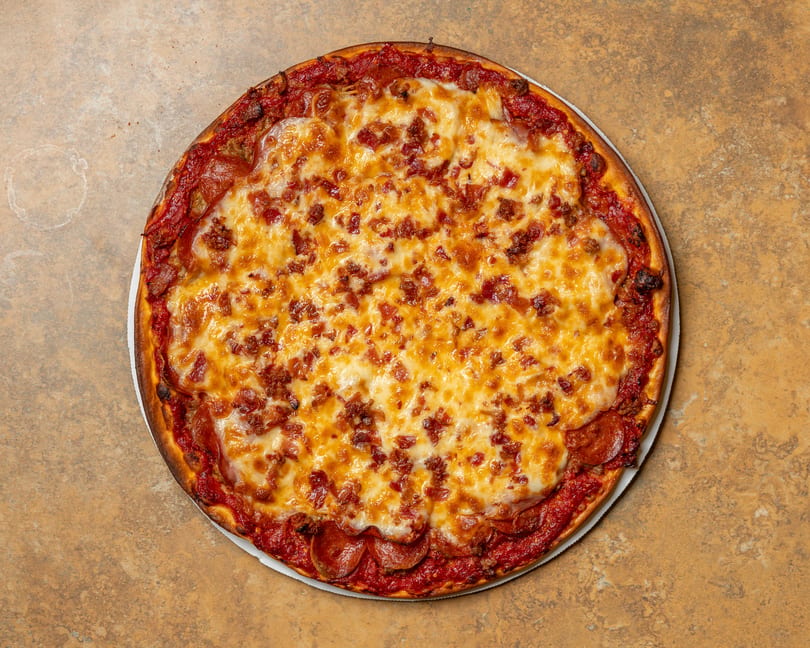 A whole pepperoni and bacon pizza with melted cheese and a thick crust on a brown surface.
