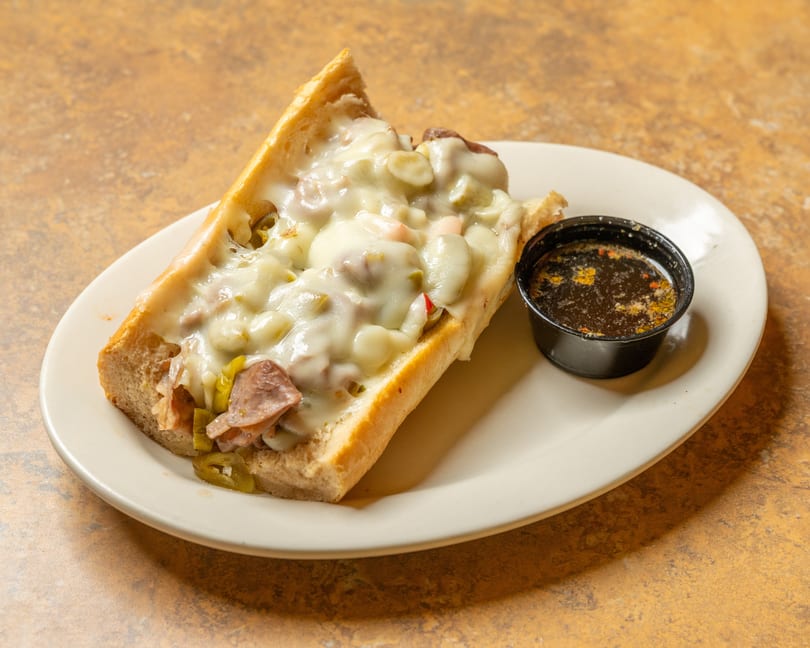 A cheesy Italian beef sandwich with giardiniera and a side of au jus on a white plate.
