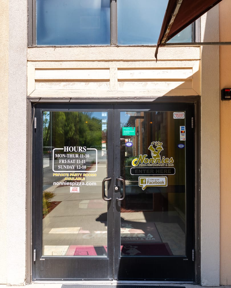 The glass entrance doors of Nonnies Pizza Restaurant & Bar, displaying hours, website, and social media.