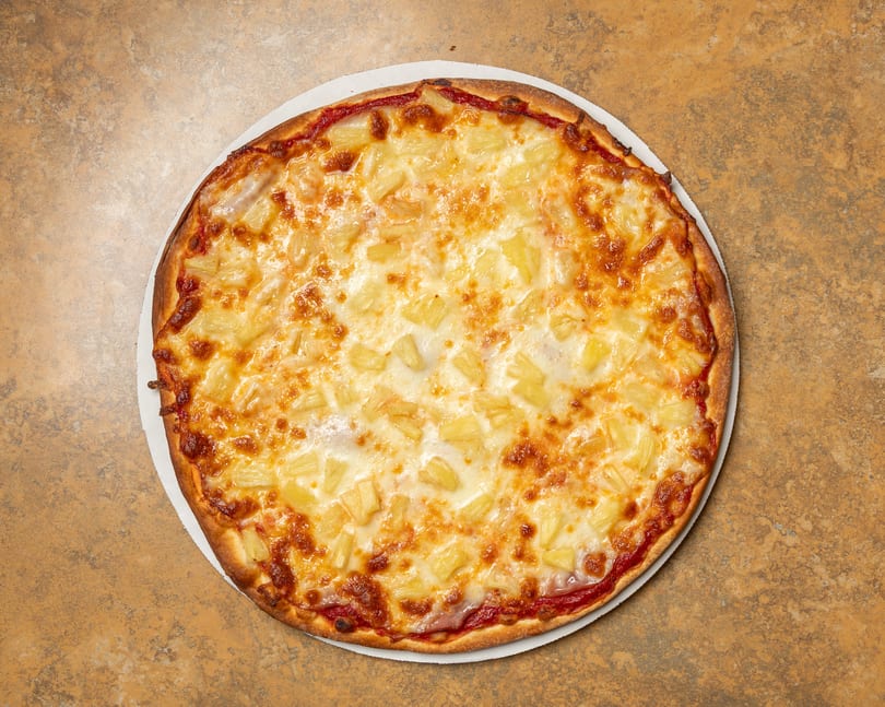 A whole pineapple pizza with melted cheese and tomato sauce on a brown textured surface.