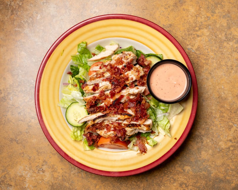 Grilled chicken salad with bacon, lettuce, tomatoes, cucumbers, and a side of dressing.